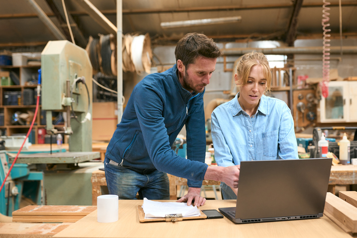 Male And Female Carpenters Working In Woodwork Workshop Using Laptop Together